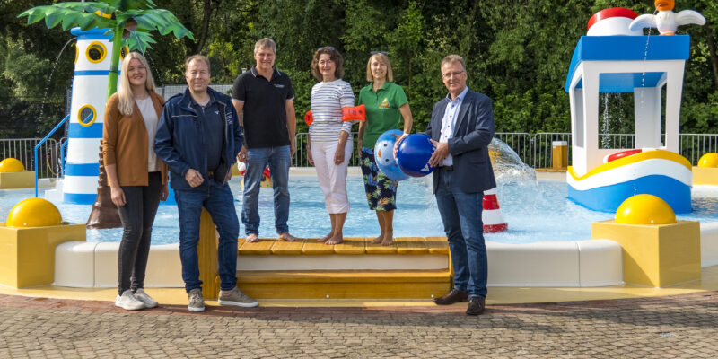 3 Damen und 3 Herren stehen am Rande eines Kinderplanschbeckens und schauen in die Kamera. Im Hintergrund sind Spritzattraktionen im Planschbecken zu sehen, darunter ein Leuchtturm, Palme, eine Boje, ein Boot mit Möwe oben drauf